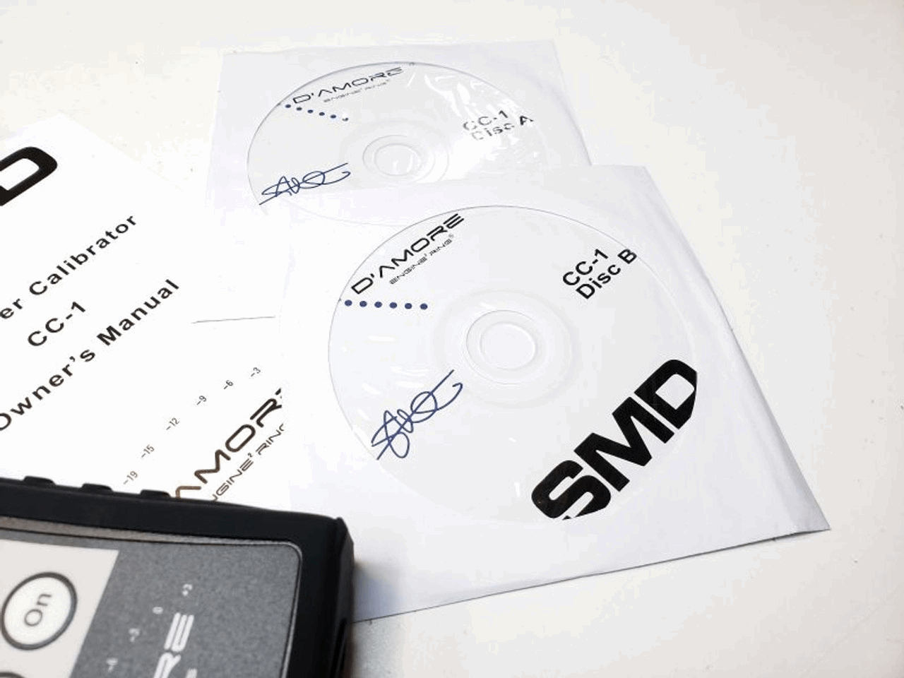 Two CDs labeled as Disc A and Disc B for the SMD Crossover Calibrator CC-1 by Steve Meade Designs are placed on a table next to sections of a user manual. Both the CDs and a device showcase the D'Amore and SMD logos, highlighting advanced gain matching for precise audio adjustment.