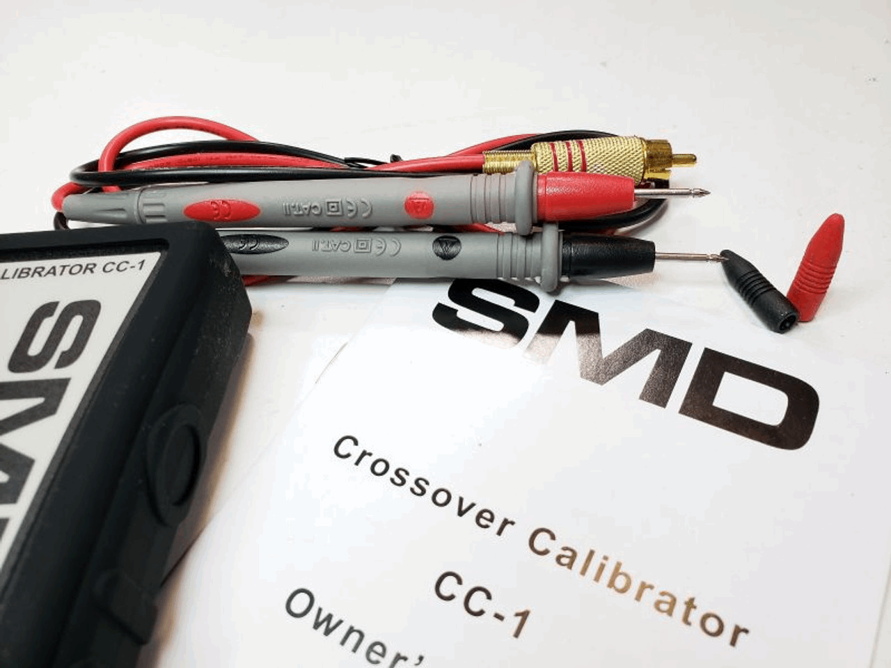 Close-up image of a pair of multimeter probes resting on a document titled SMD Crossover Calibrator CC-1 Owners. The silver probes, with red and black wires, exemplify gain matching precision. To the left, a section of a device labeled SMD Crossover Calibrator CC-1 - Steve Meade Designs is visible, hinting at its role in amplifier setups.