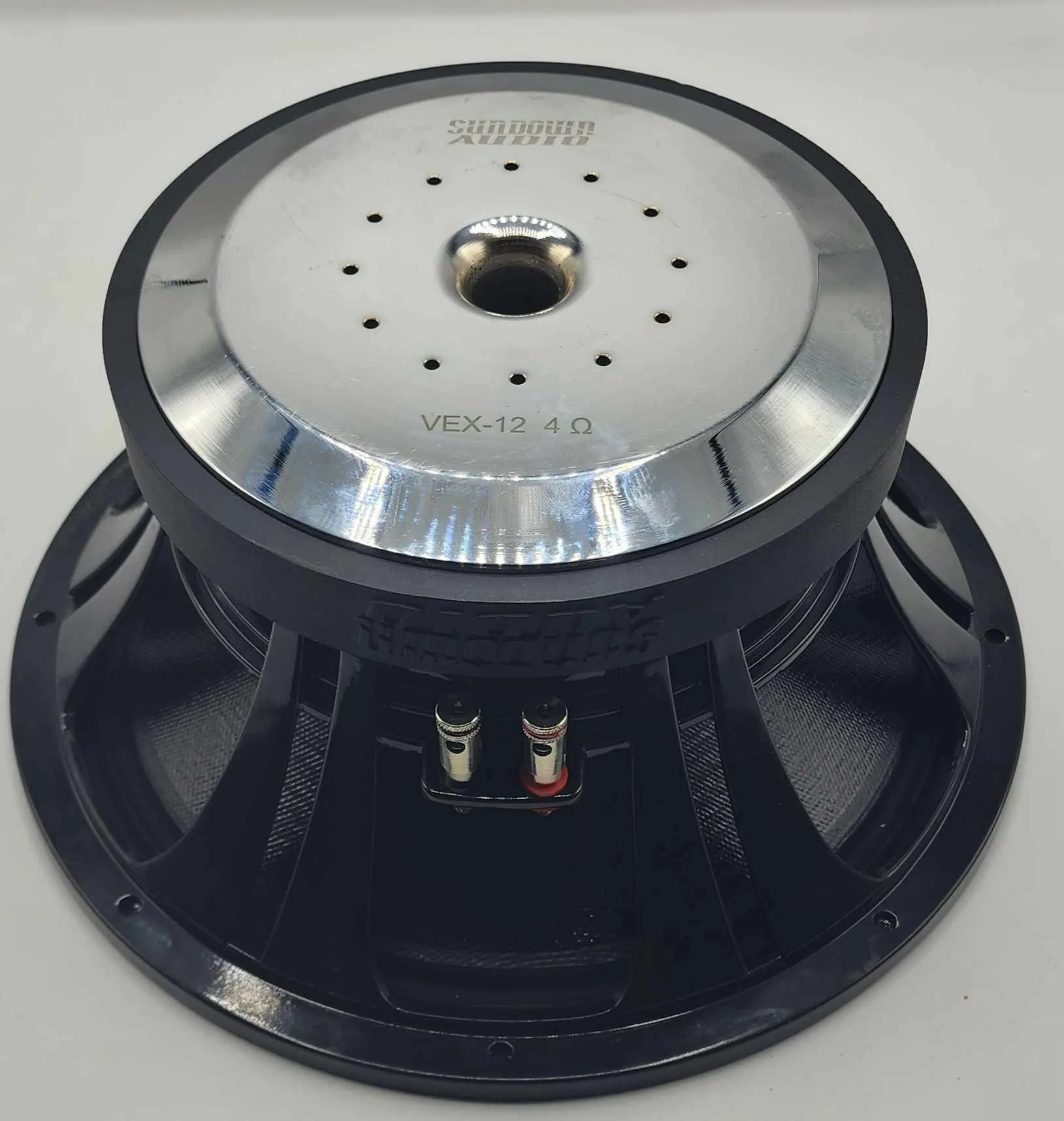 A close-up of a black and silver Sundown Audio VEX-12 500W 12" Pro Audio Midrange Speaker highlights its high-efficiency mid-range with a carbon-fiber cone. The metallic center features several small holes, and it's mounted on a black frame with two red and black terminals at the back.